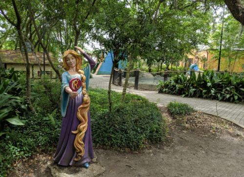A colorful statue of a princess with long hair stands in a lush garden, surrounded by trees and pathways.