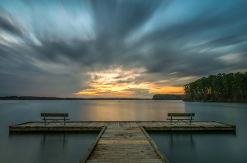 A serene lake at sunset, featuring a wooden dock with two benches and dramatic clouds in the sky.