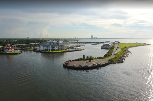 Aerial view of a marina with boats, a grassy peninsula, and a road lined with cars along the water's edge.