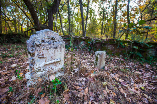 An old, weathered gravestone surrounded by overgrown grass and fallen leaves in a wooded area.