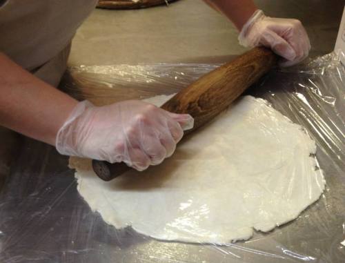 A person in gloves rolls out dough with a wooden rolling pin on a plastic-covered surface.