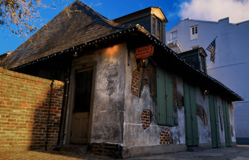 Historic building with weathered walls, green shutters, and a sign for a blacksmith shop, set against a blue sky.