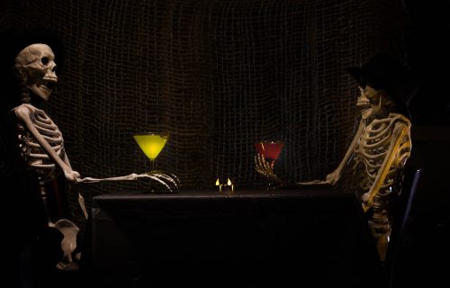 Two skeletons sit at a table with colorful cocktails, surrounded by a dark, textured backdrop.