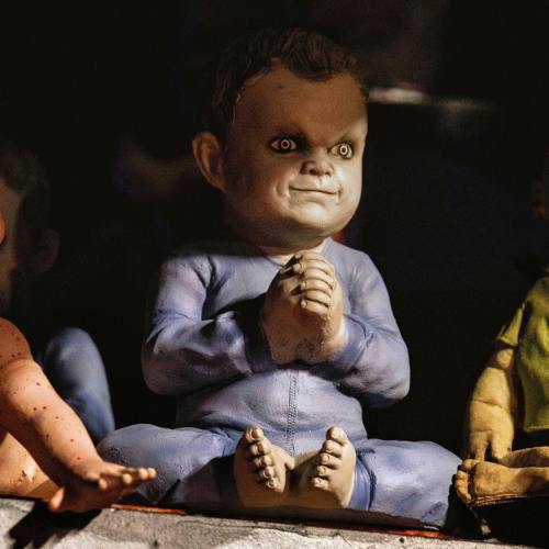 A creepy doll with a sinister smile, sitting cross-legged, wearing a blue outfit and having unsettling eyes.