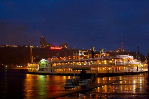 A lit-up boat docked at night, with city lights reflecting on the water in the background.