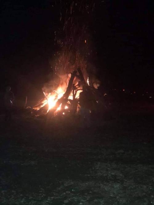 A large bonfire burns brightly at night, with sparks flying into the dark sky. Silhouettes of people are nearby.
