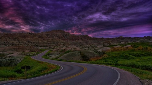 A winding road through colorful hills under a dramatic purple and blue sky at dusk.