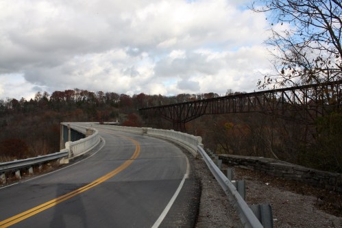 A winding road curves towards a bridge, surrounded by trees with autumn foliage under a cloudy sky.