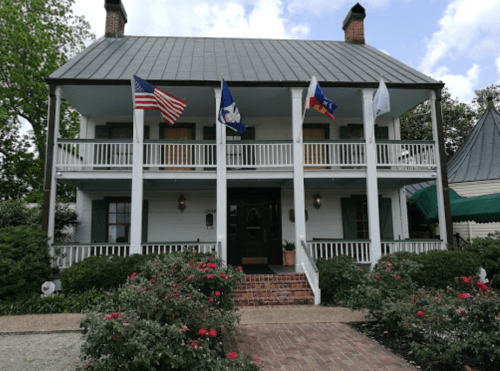 Historic two-story house with flags, green shutters, and a porch, surrounded by blooming rose bushes.