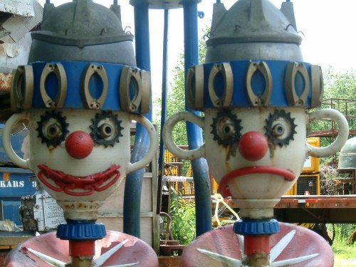 Two whimsical clown figures with gear eyes and painted expressions, one smiling and one frowning, set against a rustic background.