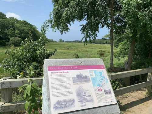 A scenic view of Namasket Creek with a sign about the Cape Cod Rail Trail in the foreground, surrounded by greenery.