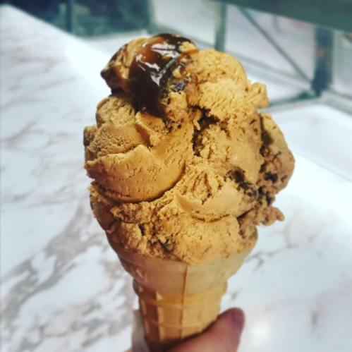 A hand holding a cone of caramel-flavored ice cream topped with a drizzle of caramel sauce.