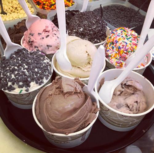 A tray of eight ice cream cups in various flavors, topped with sprinkles and chocolate chips, with spoons in each cup.