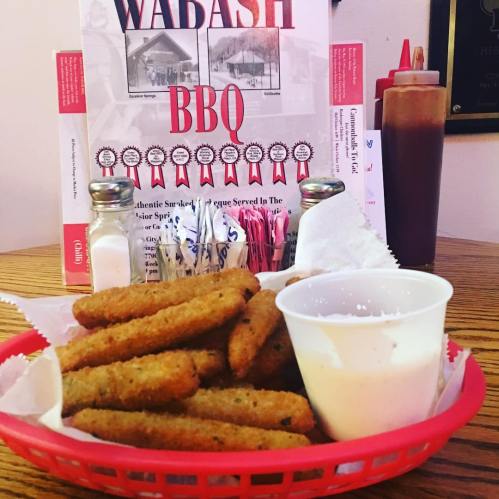 A basket of fried pickles with a side of dipping sauce, set against a BBQ restaurant backdrop.