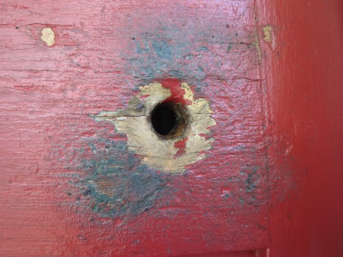 Close-up of a weathered red wooden surface with a circular hole, showing peeling paint and textured wood grain.
