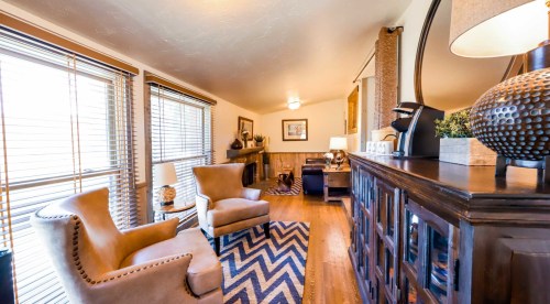 Cozy living room with two armchairs, a patterned rug, and large windows with blinds, featuring warm decor and lighting.
