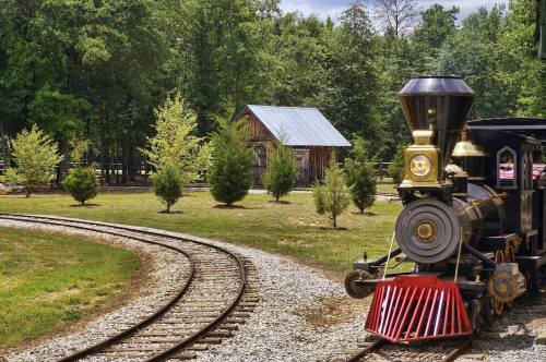 A vintage steam train on curved tracks near a rustic barn and green trees in a scenic outdoor setting.