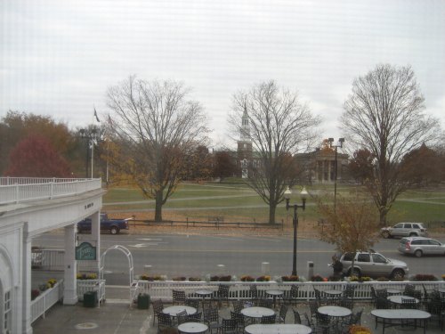 View of a park with bare trees and autumn foliage, featuring a clock tower in the background and outdoor seating in the foreground.