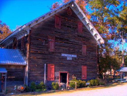 Historic Kymulga Mill, a weathered wooden building with red shutters, surrounded by trees and a clear blue sky.