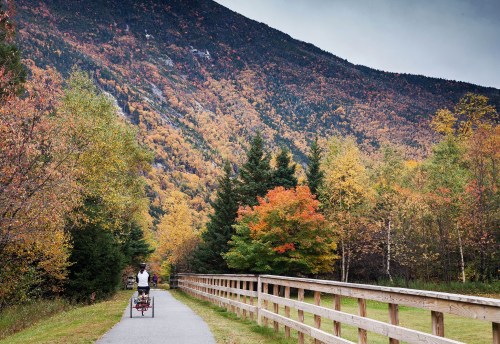 A cyclist rides along a path surrounded by vibrant autumn foliage and mountains in the background.