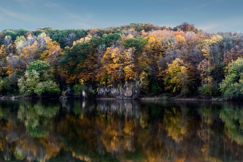 A serene river scene with autumn-colored trees reflecting on calm water under a clear sky.