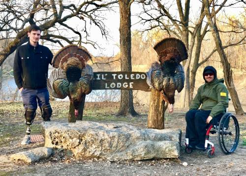 Two men pose beside a wooden sign for "Il Toledo Lodge," flanked by two decorative turkeys in a wooded area.