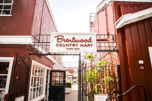 Sign for Brentwood Country Mart, established in 1948, framed by rustic buildings and greenery.