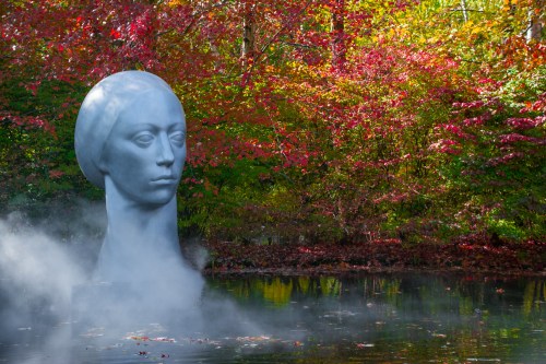 A large, serene sculpture of a woman's head rises from a misty pond, surrounded by vibrant autumn foliage.