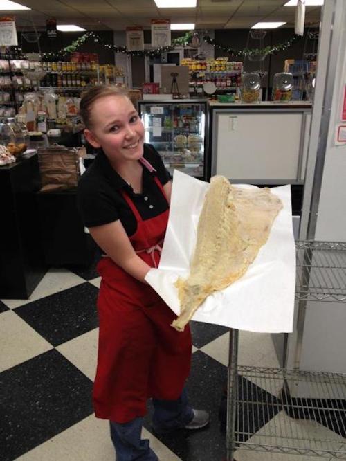 A smiling woman in an apron holds a large piece of fish on a paper towel in a grocery store.