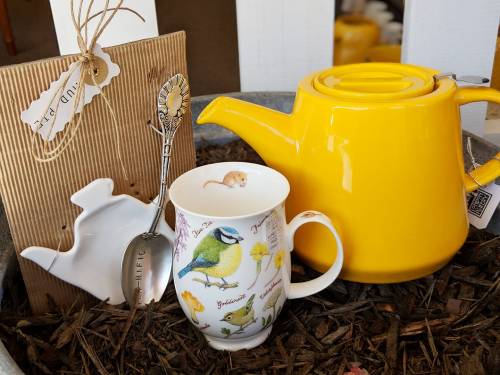 A yellow teapot, a decorative spoon, and a bird-themed mug arranged in a rustic container with mulch.