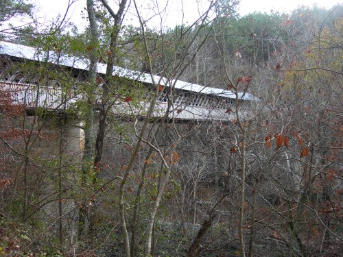 A wooden bridge partially obscured by bare trees and underbrush in a wooded area.