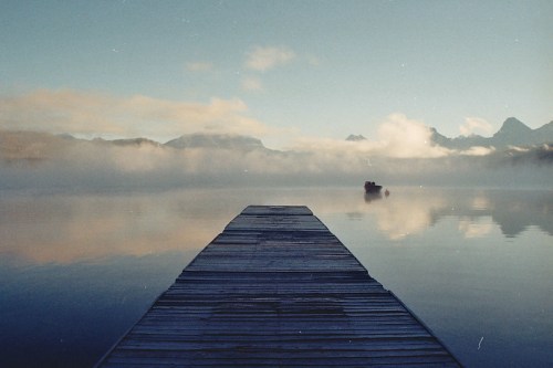 A wooden dock extends into a calm lake, surrounded by misty mountains and a serene sky.
