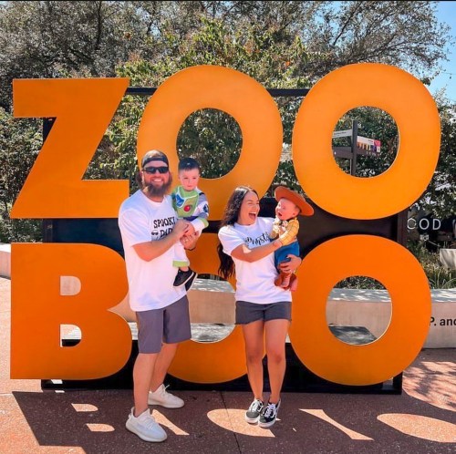 A family poses with two young children in front of a large "ZOO BOO" sign, smiling and enjoying a sunny day.