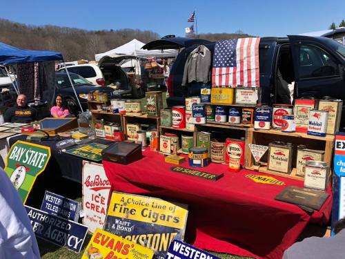 A vibrant outdoor market scene featuring vintage signs and cans on display, with people sitting behind the booth.