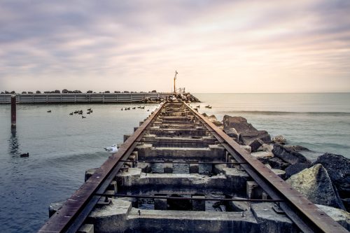 A weathered pier extends into calm waters, with ducks swimming nearby and a cloudy sky above.