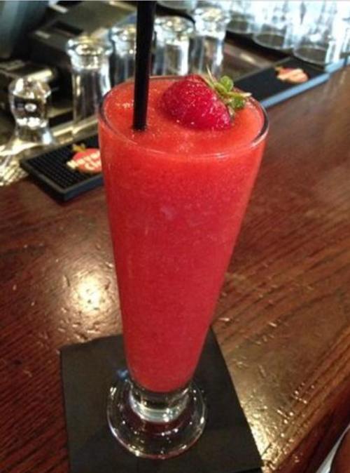 A tall glass of vibrant red slushy drink garnished with a strawberry, resting on a black napkin at a bar.