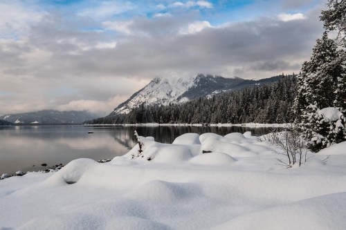A serene winter landscape featuring snow-covered hills, a calm lake, and a backdrop of mountains under a cloudy sky.