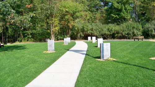 A winding concrete path through a grassy area, lined with stone pillars and surrounded by trees.