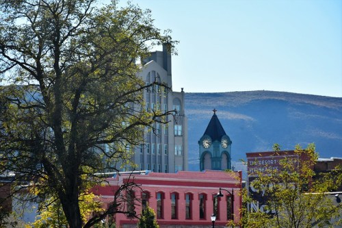 A cityscape featuring a clock tower, modern buildings, and a mountain backdrop, framed by tree branches.