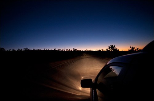 A car's headlights illuminate a dirt road at dusk, with a gradient of blue and orange in the sky and silhouettes of trees.