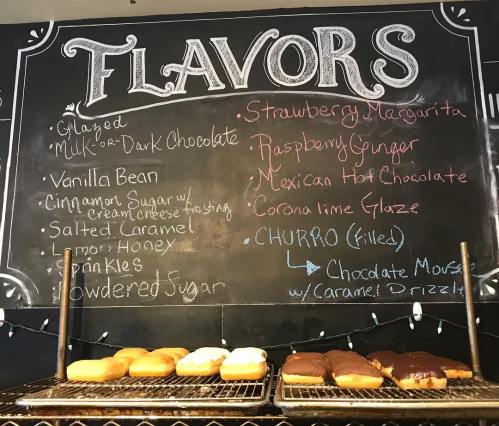 Chalkboard menu listing various donut flavors, including Strawberry Margarita and Churro, with donuts displayed below.