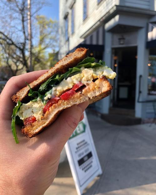 A hand holding a sandwich with greens, tomatoes, and a creamy filling, in front of a café.