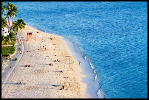 Aerial view of a sandy beach with people sunbathing, palm trees, and a lifeguard tower by the blue ocean.