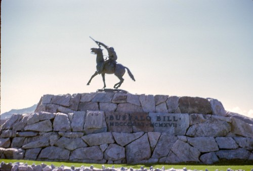 Statue of Buffalo Bill on a stone pedestal, with a horse and rider, against a clear blue sky.