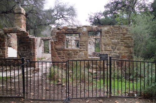 Ruins of a stone building surrounded by a black fence, with trees and greenery in the background.