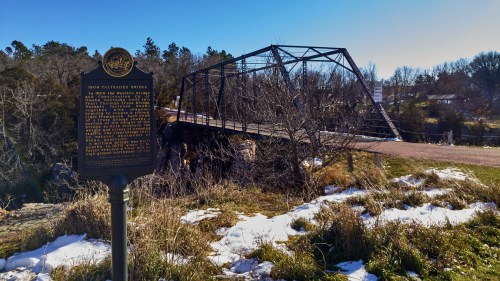Historic sign in foreground with the 1908 Palisades Bridge in the background, surrounded by grass and snow. Clear blue sky.