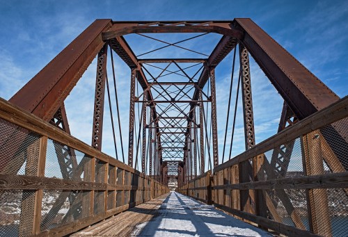 A view of a rusted metal bridge with wooden walkways, set against a clear blue sky. Snow covers the ground below.