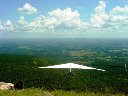 A hang glider soars over a lush green landscape under a bright blue sky with fluffy clouds.