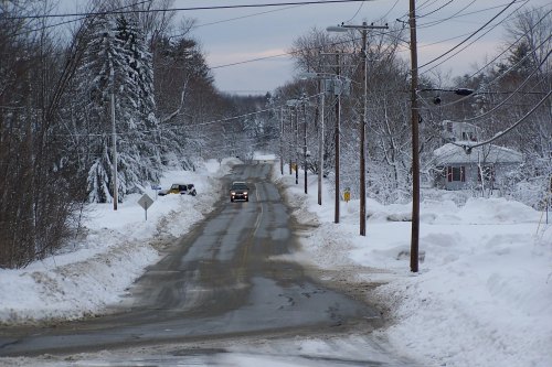 A snowy road lined with trees and power lines, with a car driving through the winter landscape.