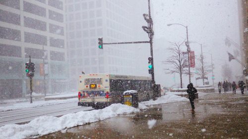 A snowy city street with a bus at a green light, pedestrians walking, and buildings partially obscured by falling snow.